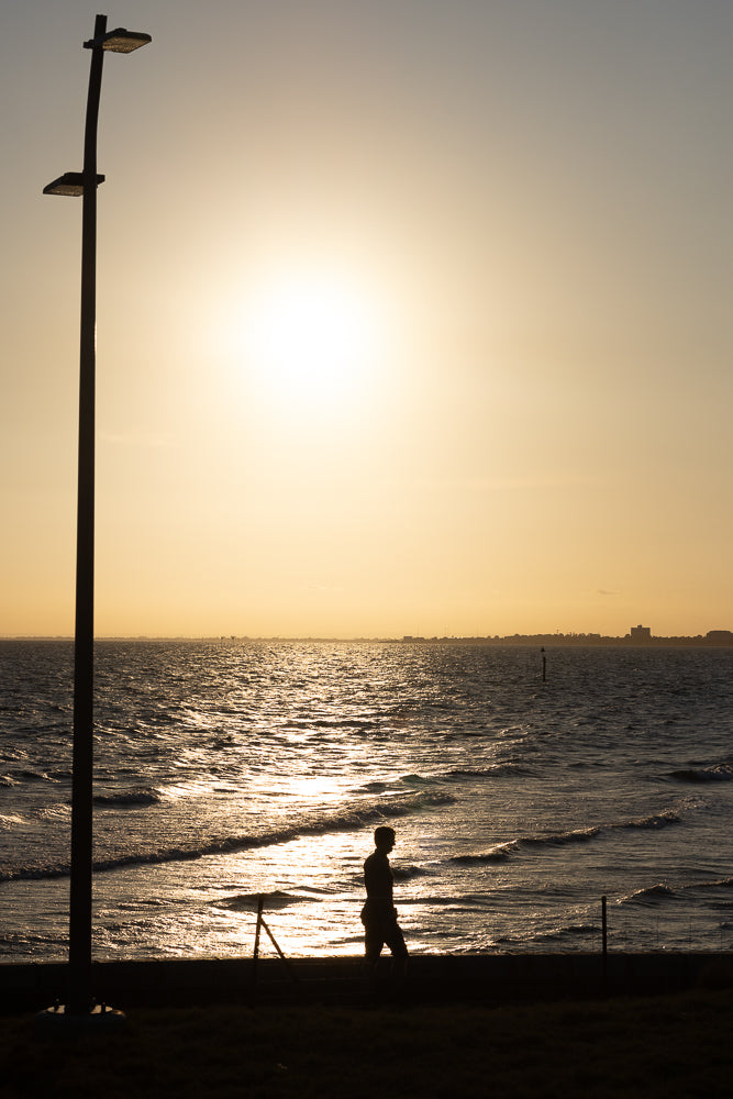 A silhouette of a person walks along the shore as the sun sets over the ocean. The sun reflects off the water, creating a shimmering path. A tall lamppost stands to the left of the frame.