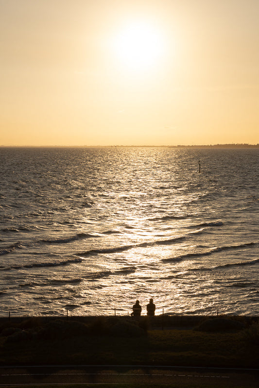 Two people silhouetted against a bright, hazy sunset over the ocean. The sun's reflection creates a shimmering path across the water. They sit on a grassy embankment overlooking the sea.