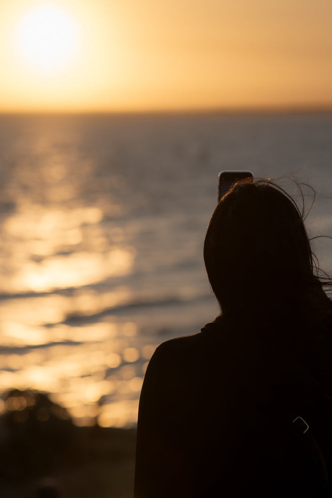 A person in silhouette is taking a photo of a sunset over the ocean with their phone.