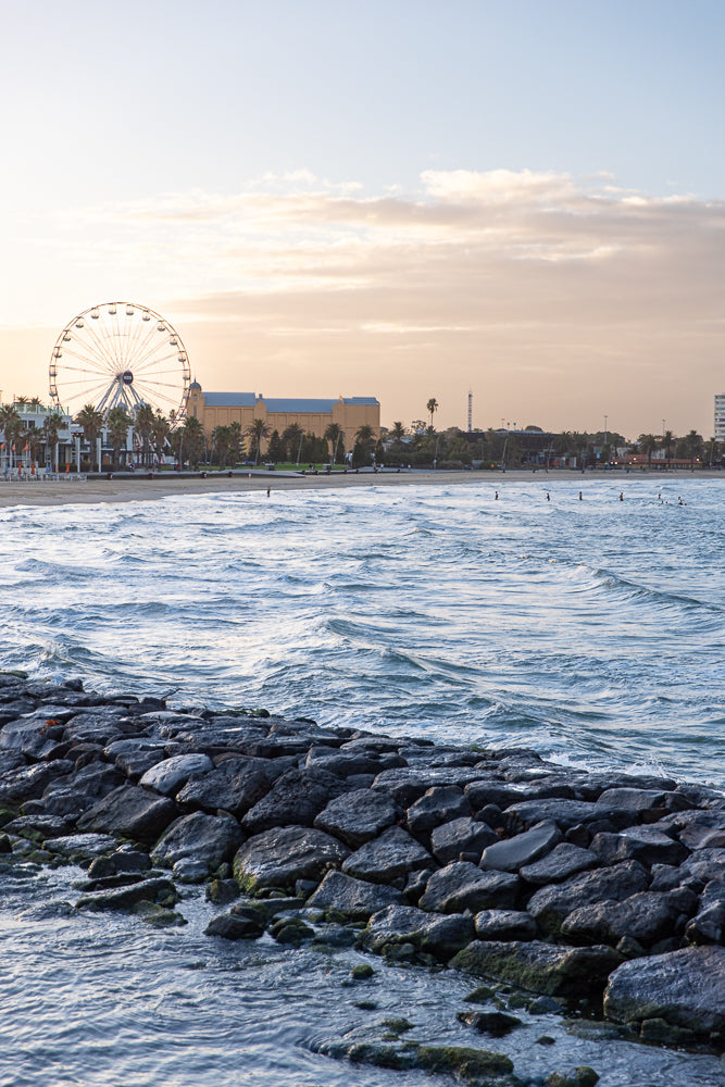 A wide shot of a beach scene at dusk. A large Ferris wheel dominates the skyline, with palm trees and buildings lining the shore. The ocean has gentle waves, and a rocky breakwater is in the foreground.
