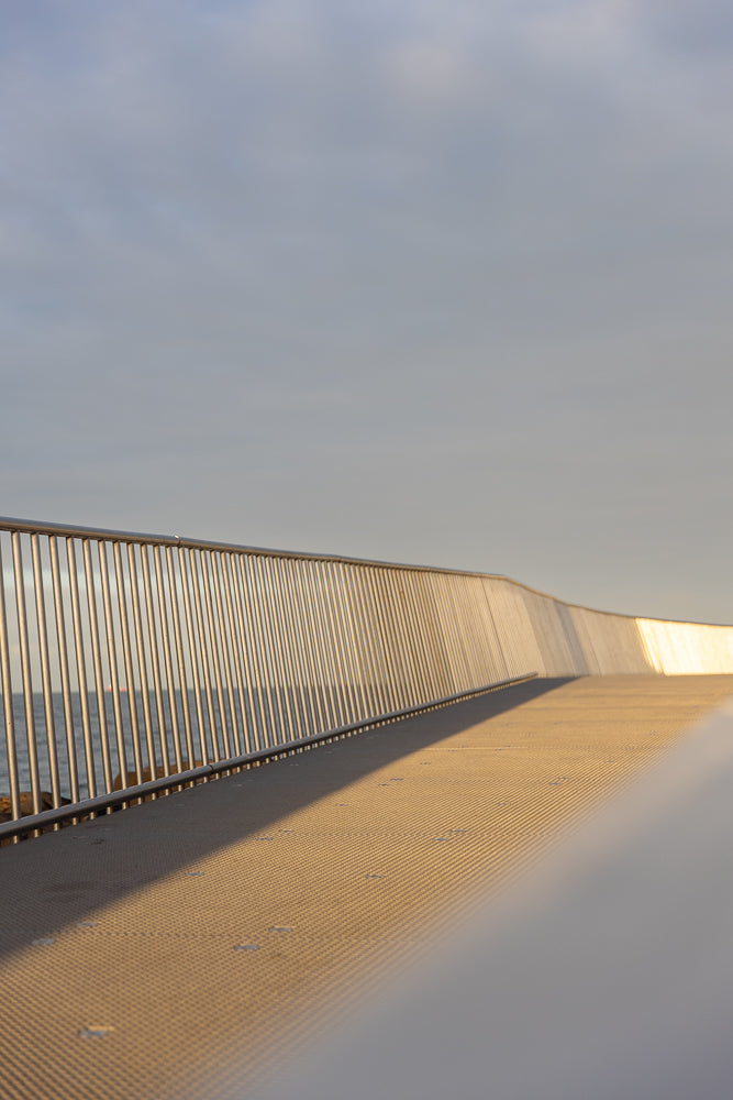 A modern metal railing and walkway curve along the edge of the water under a pale sky. The walkway is illuminated by warm, low sunlight, casting long shadows.