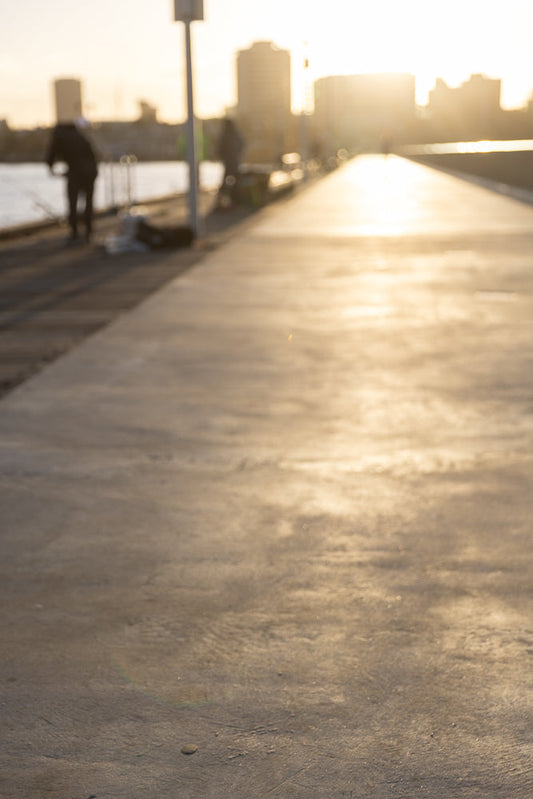 A person walks along a paved path next to water as the sun sets, casting a warm glow on the scene. Buildings are visible in the distance.