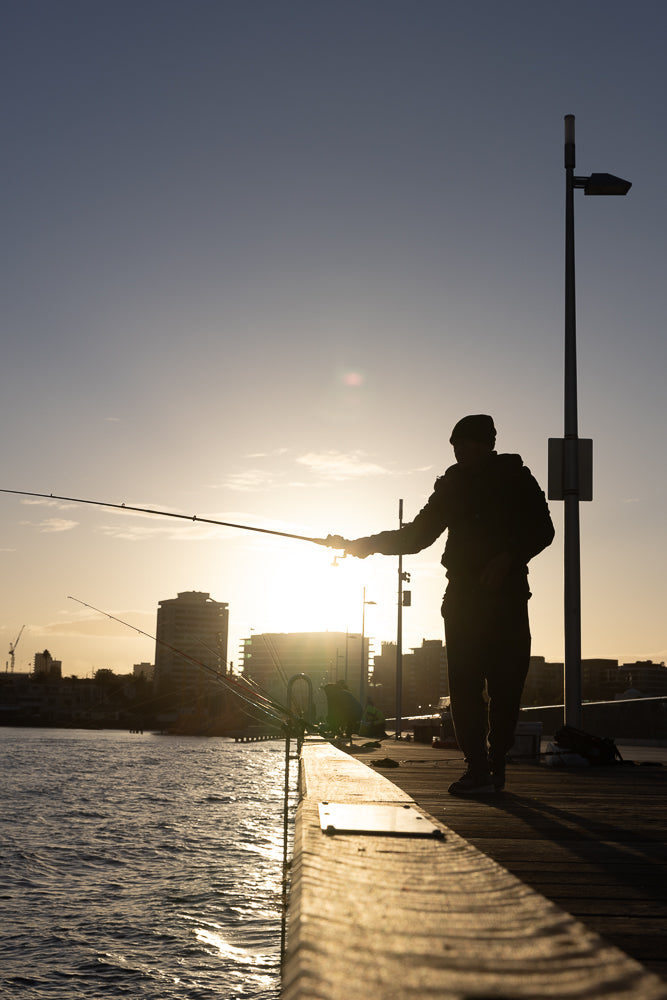 Silhouette of a person fishing on a pier at sunset. The sun is low on the horizon, casting a warm glow and long shadows. Buildings and a crane are visible in the background across the water.