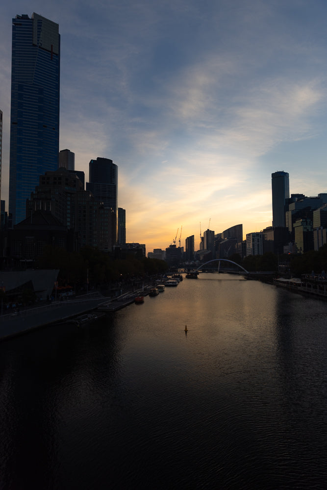 A city skyline at dusk with a river in the foreground. Tall buildings and cranes are visible against a sky with warm sunset colors and clouds.