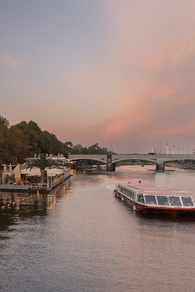 A wide shot of a river at dusk with a large passenger boat moving away from the camera. On the left, a floating restaurant with people dining under umbrellas and string lights is visible. A bridge spans the river in the background, with a soft pink and purple sky above.