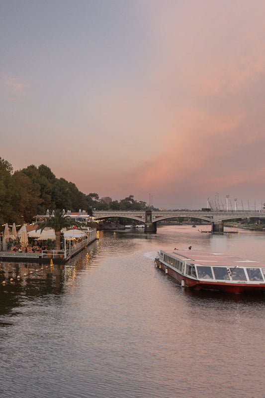 A wide shot of a river at dusk with a large passenger boat moving away from the camera. On the left, a floating restaurant with people dining under umbrellas and string lights is visible. A bridge spans the river in the background, with a soft pink and purple sky above.