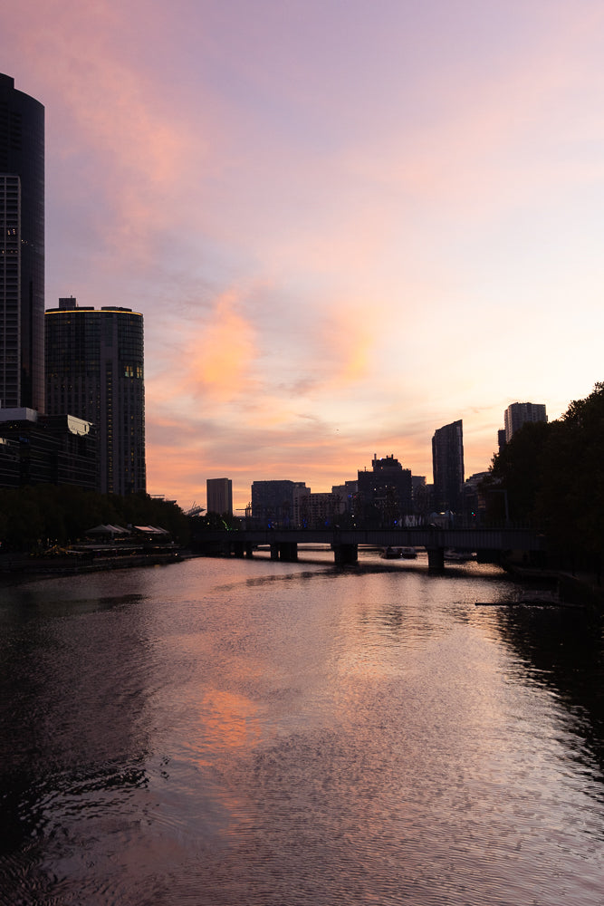 A city skyline at sunset with a river in the foreground. The sky is filled with soft pink and orange clouds, reflecting on the water. Buildings and a bridge are visible in the background.