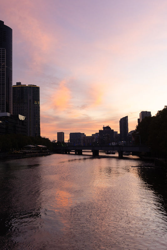 A city skyline at sunset with a river in the foreground. The sky is filled with soft pink and orange clouds, reflecting on the water. Buildings and a bridge are visible in the background.