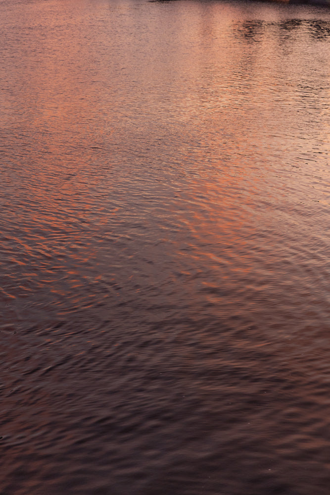 The surface of water reflects the warm, orange and pink hues of a sunset. Small ripples create a textured pattern across the water.