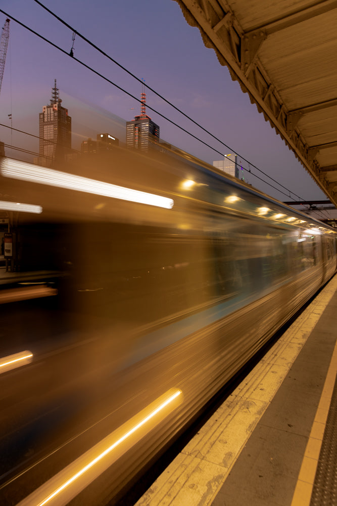 A train speeds through a station at dusk, blurring past the platform as city lights twinkle in the background.