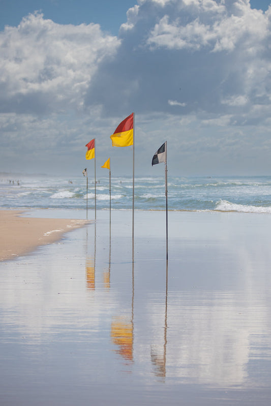 A line of surf lifesaving flags, including red and yellow, yellow, and black and white checkered flags, stand on a wet sandy beach. The flags are reflected in the shallow water covering the sand. In the background, gentle waves roll onto the shore under a cloudy sky.