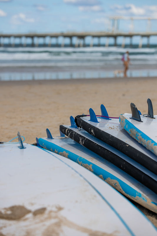 A stack of surfboards with blue and black fins rests on a sandy beach. In the background, a pier stretches into the ocean under a partly cloudy sky, and a person walks along the shoreline.