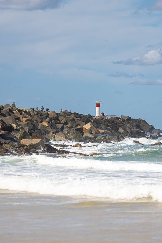 A lighthouse with red and white stripes stands on a rocky breakwater. Several people are scattered along the rocks, some looking out at the ocean. Waves crash against the rocks and the shore in the foreground.