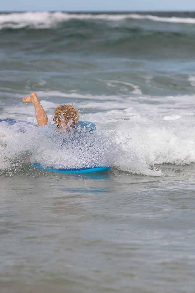 A young boy with curly blonde hair is riding a blue boogie board through a wave at the beach. His left leg is kicked up in the air as he is covered in white spray from the wave.