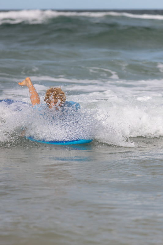 A young boy with curly blonde hair is riding a blue boogie board through a wave at the beach. His left leg is kicked up in the air as he is covered in white spray from the wave.