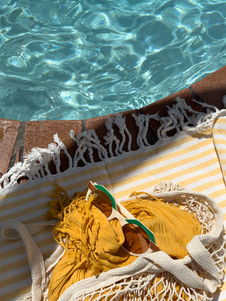 A yellow and white striped towel with a yellow scarf and sunglasses rests on the edge of a swimming pool.