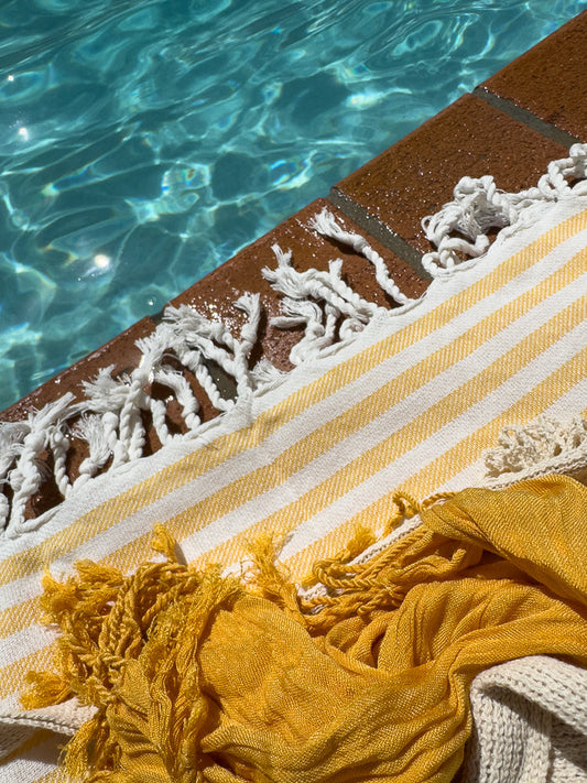 A close-up shot of a yellow and white striped towel with fringe, draped over the edge of a swimming pool. The water is a clear blue with sunlight reflecting on the surface.