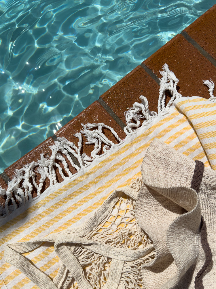 A close-up shot shows a yellow and white striped towel and a beige knit bag with fringe resting on the edge of a swimming pool. The water is clear and blue with sunlight reflecting on the surface.