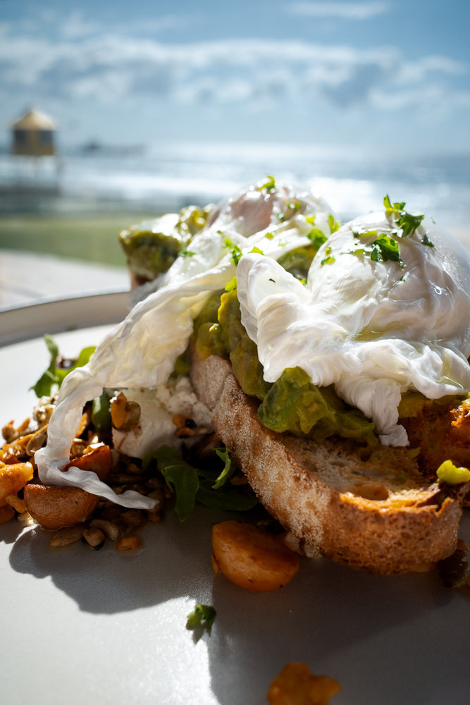 A close-up shot of avocado toast topped with poached eggs and garnished with parsley. The dish is served on a plate with a side of roasted vegetables and greens. In the background, the ocean and a lifeguard tower are visible under a bright sky.