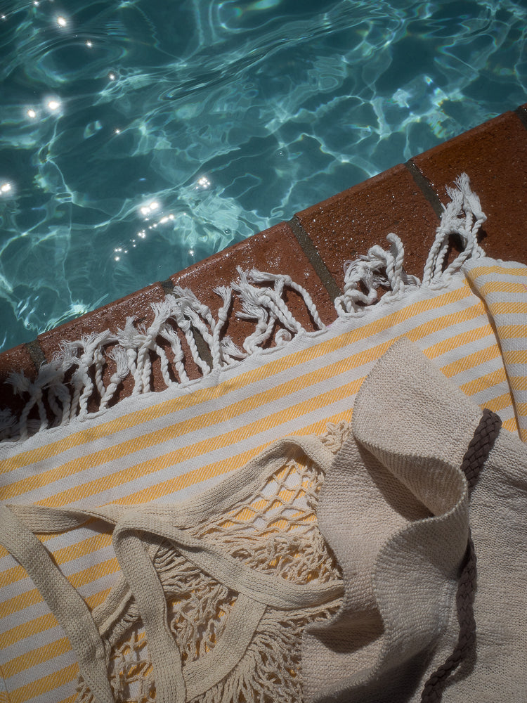 A close-up shot shows a yellow and white striped towel and a beige knitted bag with fringe, resting on the edge of a swimming pool. The water is clear blue with sunlight sparkling on the surface.