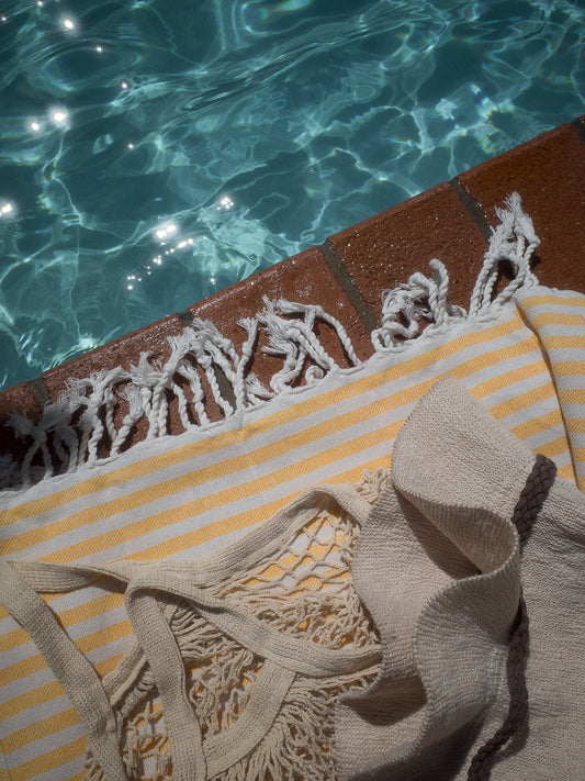 A close-up shot shows a yellow and white striped towel and a beige knitted bag with fringe, resting on the edge of a swimming pool. The water is clear blue with sunlight sparkling on the surface.