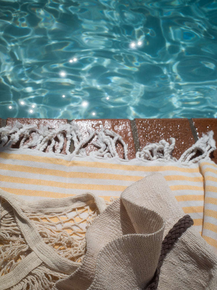 A close-up view of a striped yellow and white beach towel and a woven bag with a braided strap, laid out next to a sparkling blue swimming pool.
