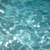 Close-up of shimmering blue water in a swimming pool, with sunlight creating bright white sparkles and abstract patterns of light and shadow on the surface.