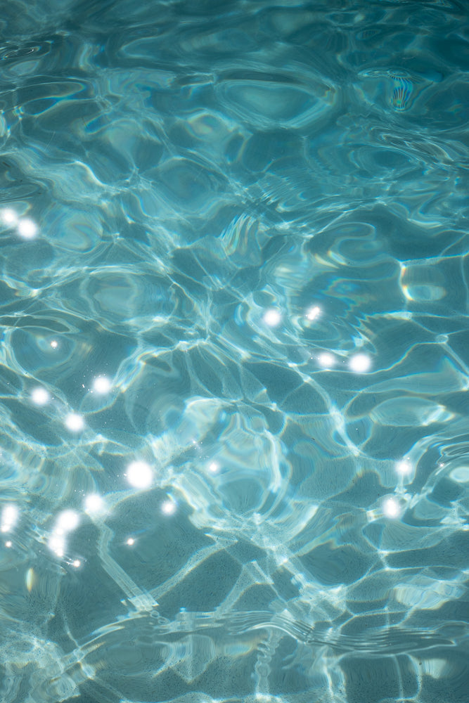 Close-up of shimmering blue water in a swimming pool, with sunlight creating bright white sparkles and abstract patterns of light and shadow on the surface.