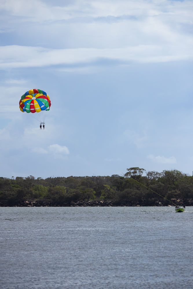 Two people parasailing in a colorful parachute over a body of water, with a tree-lined shore in the background and a boat towing them.