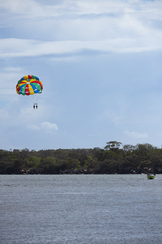Two people parasailing in a colorful parachute over a body of water, with a tree-lined shore in the background and a boat towing them.