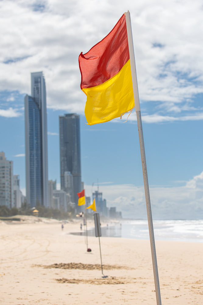 A red and yellow flag flies on a pole on a sandy beach. In the background, the ocean meets the sky under a cloudy sky. Several tall buildings are visible in the distance.
