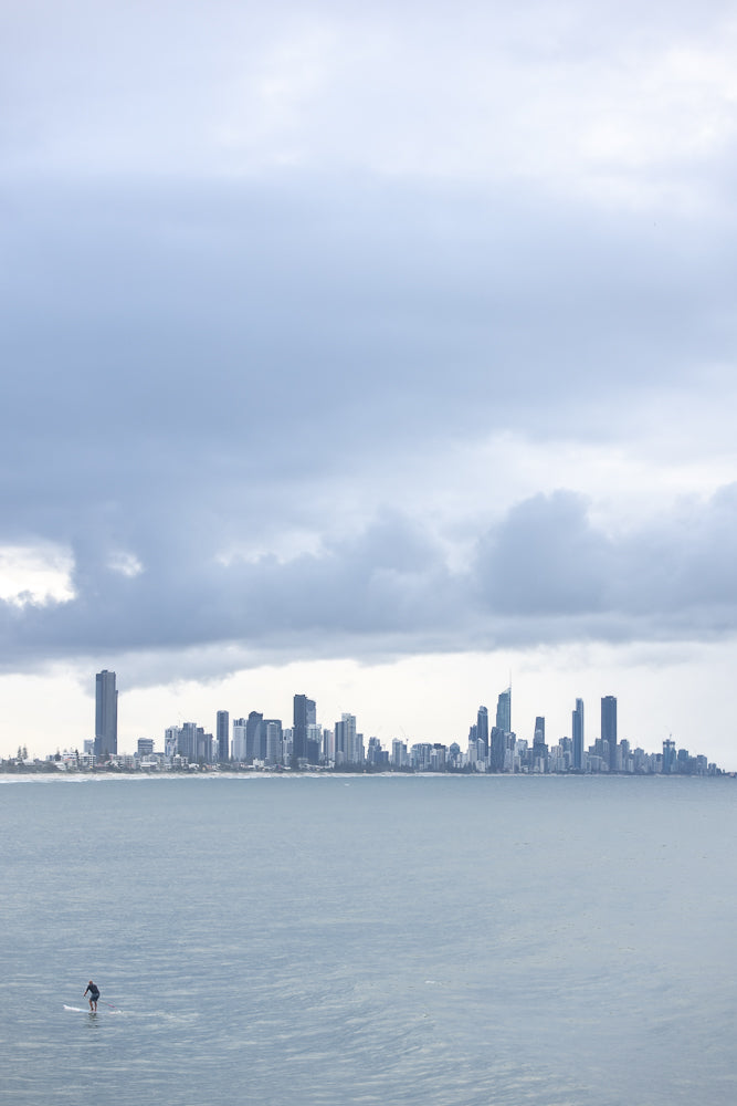 A lone paddleboarder glides across the calm ocean waters with a sprawling city skyline in the background under a cloudy sky.