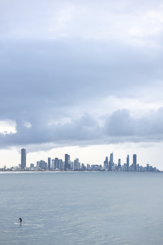 A lone paddleboarder glides across the calm ocean waters with a sprawling city skyline in the background under a cloudy sky.
