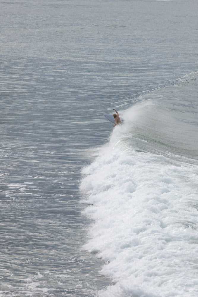 A surfer rides the crest of a wave, leaning into a turn with spray flying off the board. The surfer is shirtless and has blonde hair. The wave is breaking with white foam.