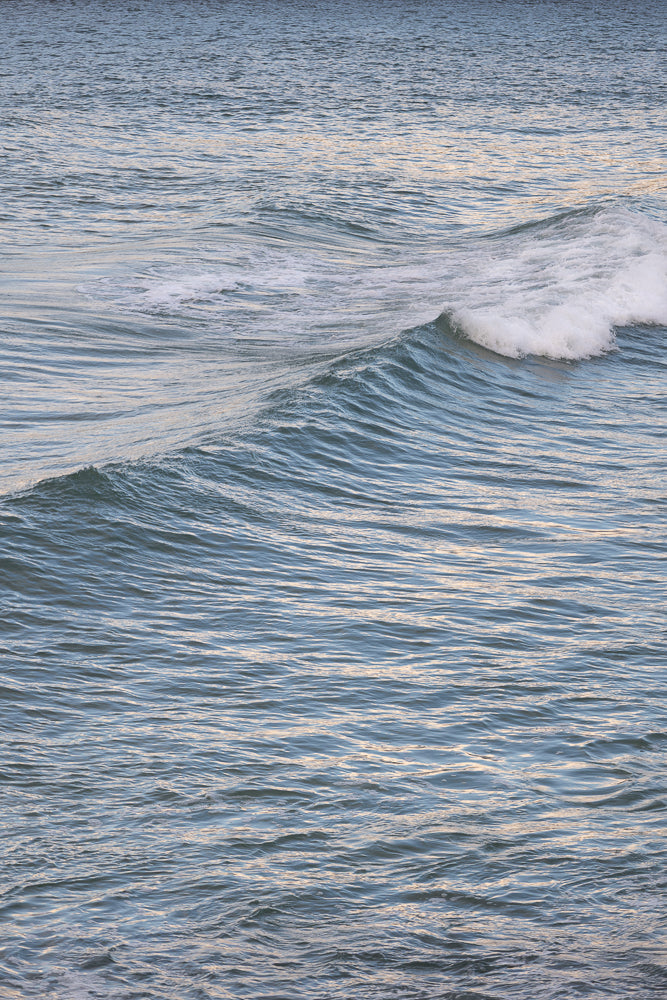 A close-up shot of the ocean surface shows gentle waves with a cresting wave on the right side. The water is a deep blue with streaks of white foam and reflects the light, creating a shimmering effect.