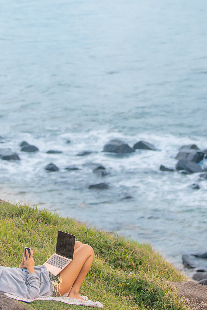A person lies on the grass near the ocean, using a laptop and phone. The ocean is in the background with waves crashing on rocks.