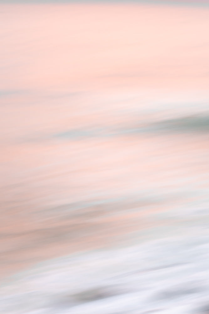 Abstract image of a beach at sunset with soft pink and peach tones blending into the pale blue of the ocean and white foam of the waves.