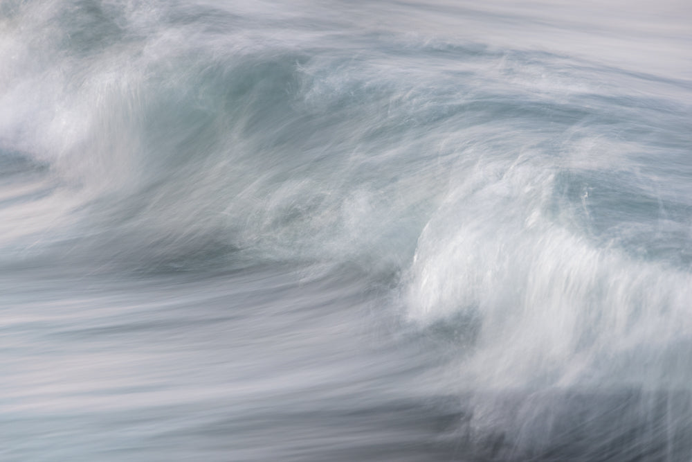 An abstract, motion-blurred image of a wave cresting and breaking. The water is a pale blue-grey, with streaks of white foam creating a sense of movement and energy.