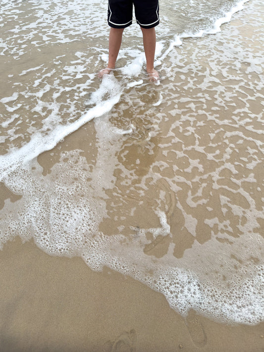 A person stands with their feet in the shallow water at the edge of the ocean. The water is clear and foamy, with small waves lapping at the sandy shore.