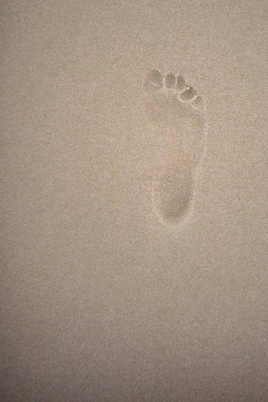 A single, clear footprint pressed into fine, light brown sand. The impression shows the shape of a bare foot, including the heel, arch, ball, and five toes.
