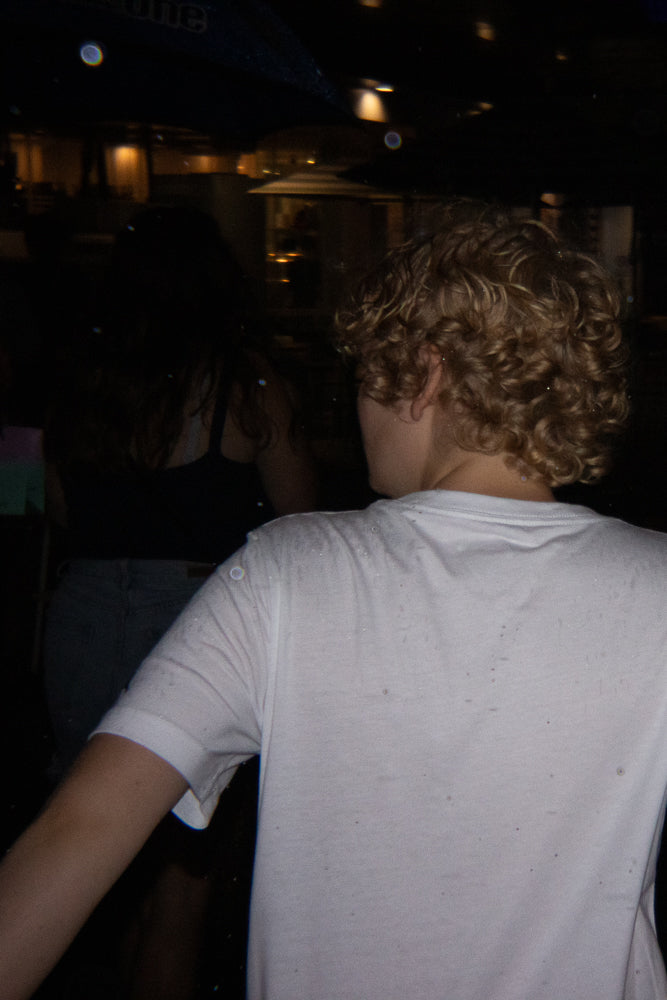 A person with curly blonde hair seen from behind, wearing a white t-shirt. The background is dark with some blurred lights and reflections.