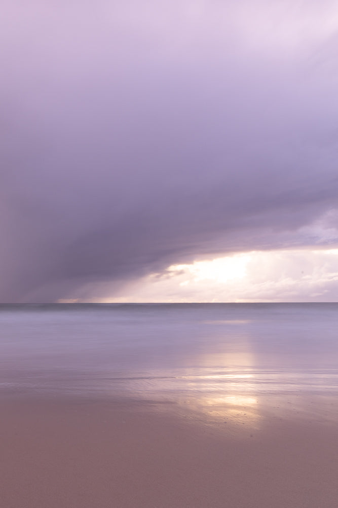 A serene, long-exposure shot of a beach at dusk. The sky is filled with soft purple clouds, with a bright golden light breaking through on the horizon. The ocean is a smooth, blurred expanse of lavender, reflecting the sky's colors. The wet sand in the foreground is also a muted purple, mirroring the gentle waves.