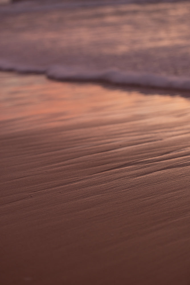 Close-up of wet sand on a beach with ripples from receding waves. The sand is a warm, reddish-brown color, and the water is a soft, muted pink and purple, suggesting a sunset or sunrise.