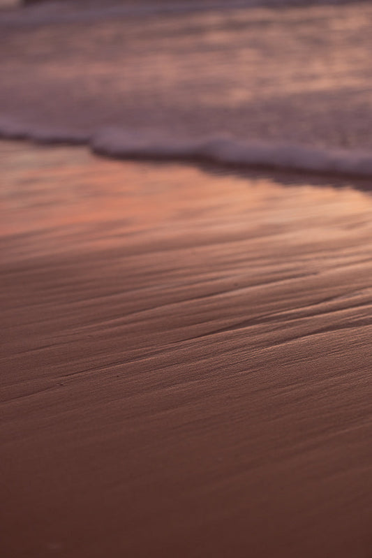 Close-up of wet sand on a beach with ripples from receding waves. The sand is a warm, reddish-brown color, and the water is a soft, muted pink and purple, suggesting a sunset or sunrise.