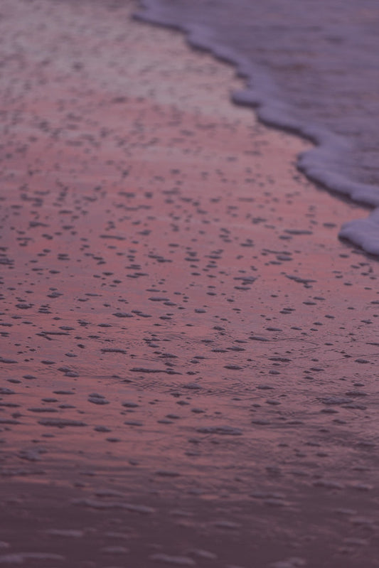 A close-up shot of the ocean's edge at sunset. The water is a soft purple hue, reflecting the sky. Small bubbles and foam from the receding tide are scattered across the wet sand.