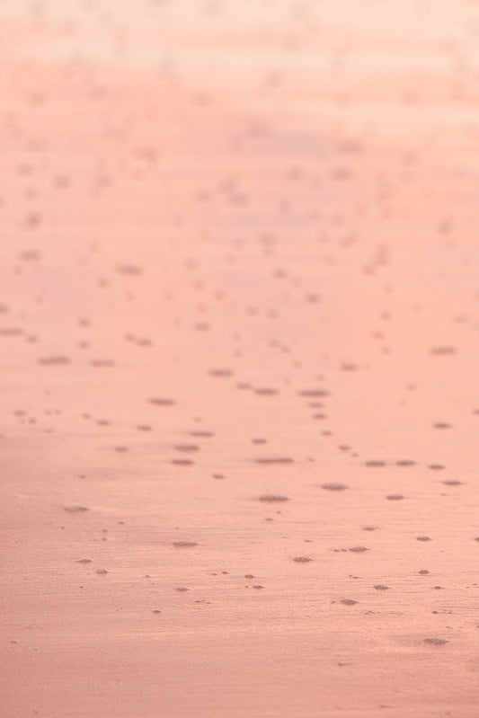 A close-up, abstract shot of wet sand with small bubbles and ripples, illuminated by soft, warm light, creating a serene and textured background.
