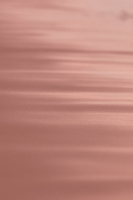 A soft focus, abstract background with horizontal lines of light and shadow in shades of rose gold and pink. The texture appears slightly grainy, creating a subtle, shimmering effect.
