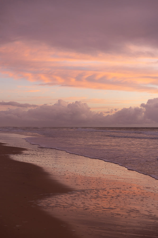 A serene beach scene at sunset, with soft pink and purple clouds reflecting on the wet sand and gentle waves rolling ashore.