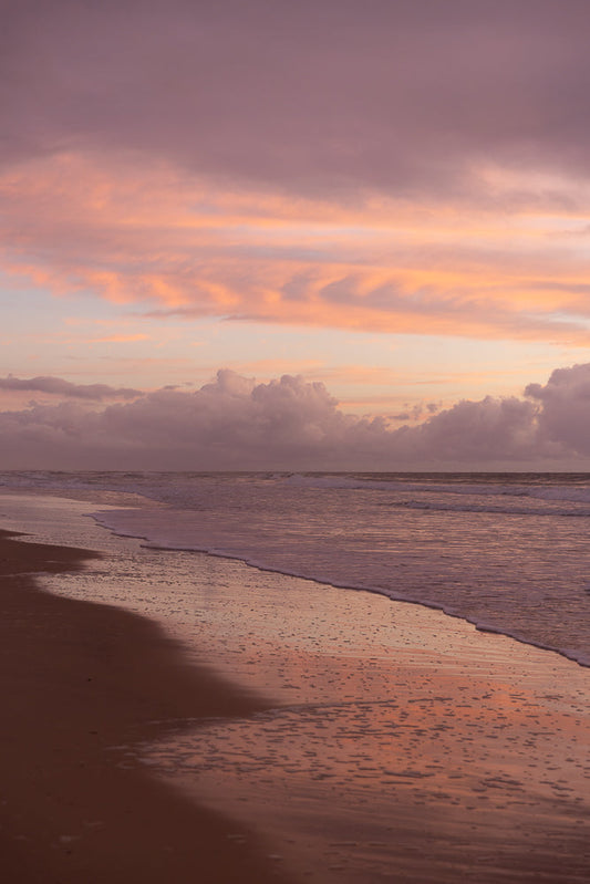 A serene beach scene at sunset, with soft pink and purple clouds reflecting on the wet sand and gentle waves rolling ashore.