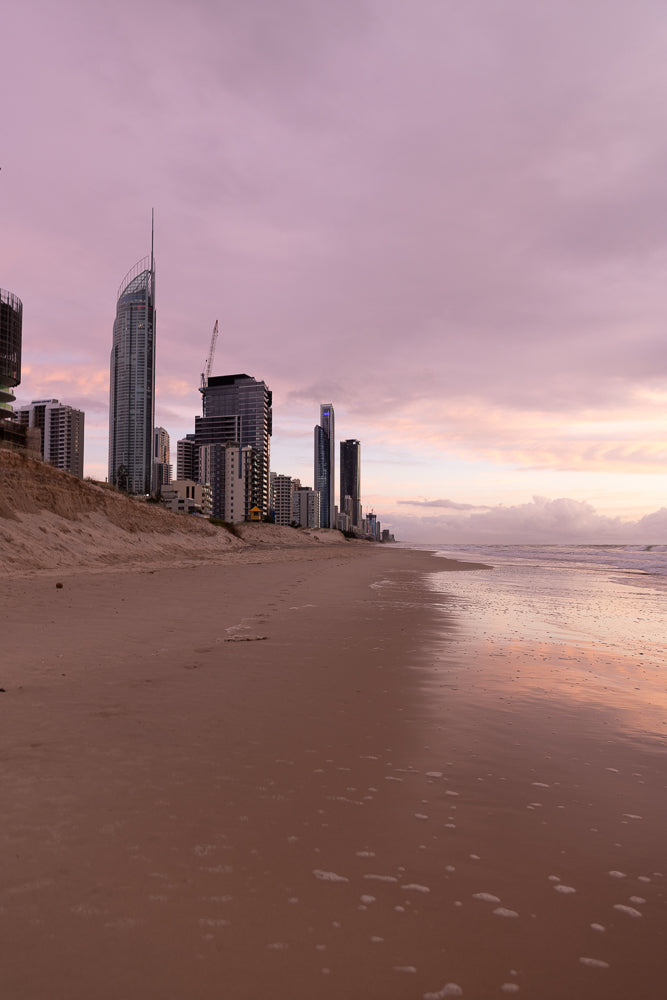 A wide shot of a beach with a city skyline in the background during a pink and purple sunrise. The ocean waves gently lap the shore, reflecting the soft colors of the sky. The sand is wet and smooth, with some footprints visible.